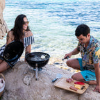 Friends grilling on the Napoleon NK14 Portable Charcoal Kettle on seaside rocks